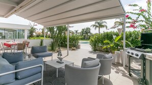 Un patio exterior con zona de estar cubierta, una parrilla y una piscina al fondo.