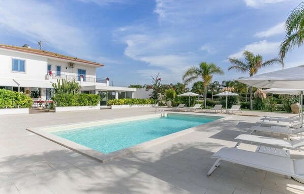 A swimming pool with lounge chairs and umbrellas, surrounded by palm trees and a building with blue shutters. - Beach front apartment in Marina di Ragusa (Marina di Ragusa)