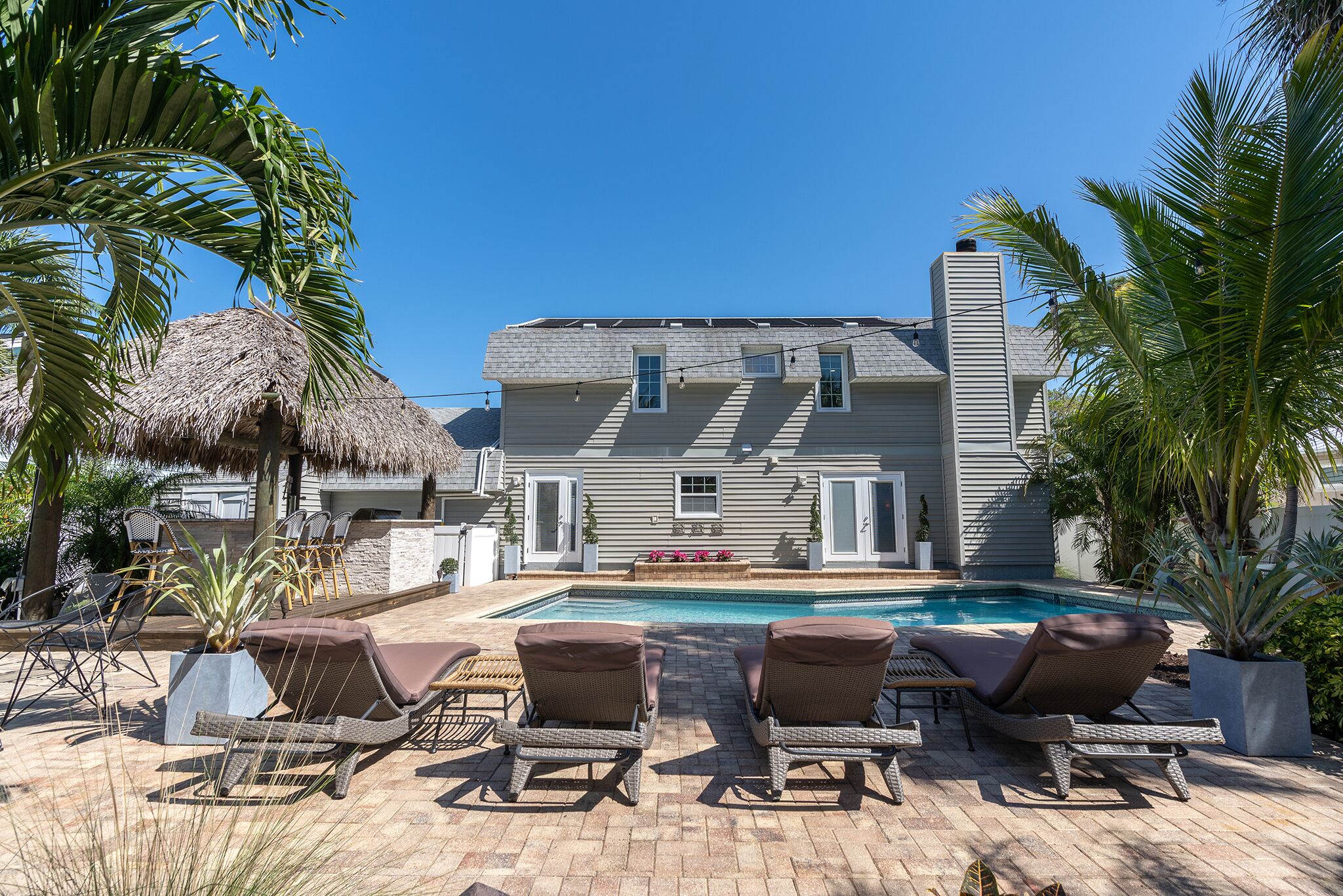Une zone de piscine avec des chaises longues, une structure à toit de chaume et une maison en arrière-plan.