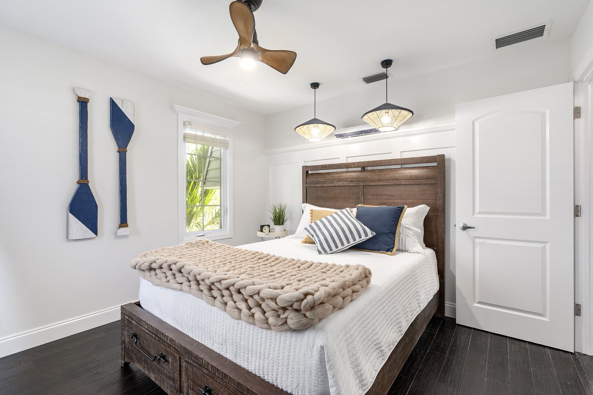 Une chambre dotée d’un grand lit, d’une tête de lit en bois, d’un ventilateur de plafond, de luminaires suspendus et d’une fenêtre donnant sur un paysage verdoyant.