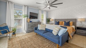 A modern bedroom with a bed, a sofa, a TV, and a ceiling fan. - Coastal Vibe • Private Heated Pool • Hot Tub • Near Beaches (Clearwater)
