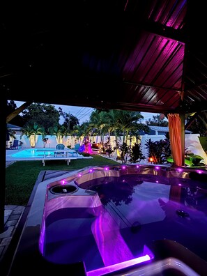 Un jacuzzi con iluminación púrpura, una piscina y mobiliario exterior.