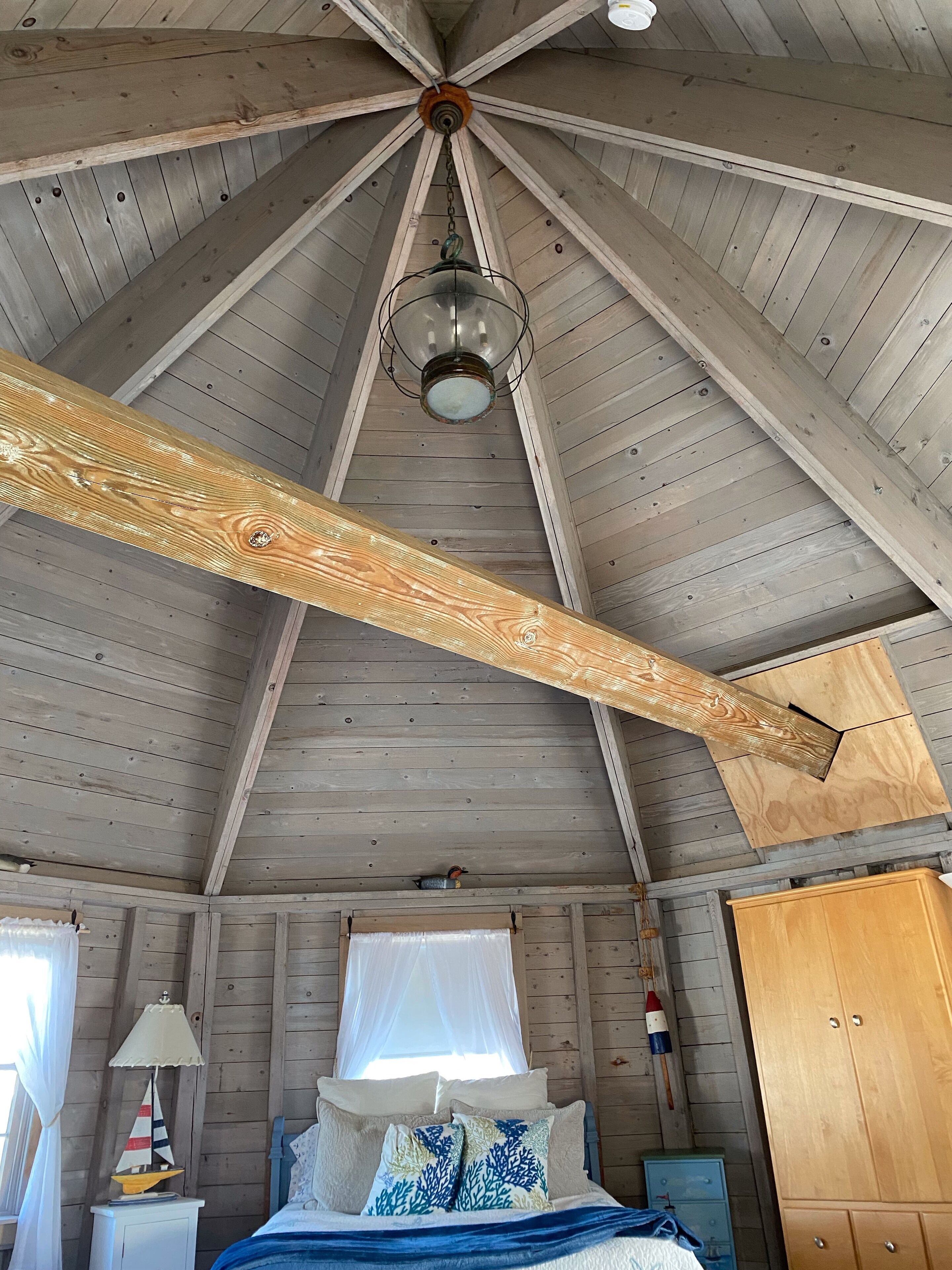 Eine Hütte mit geneigtem Dach, einem Bett mit Kissen, einem Nachttisch, einer Lampe, einem Kleiderschrank und einem Fenster mit Vorhang.