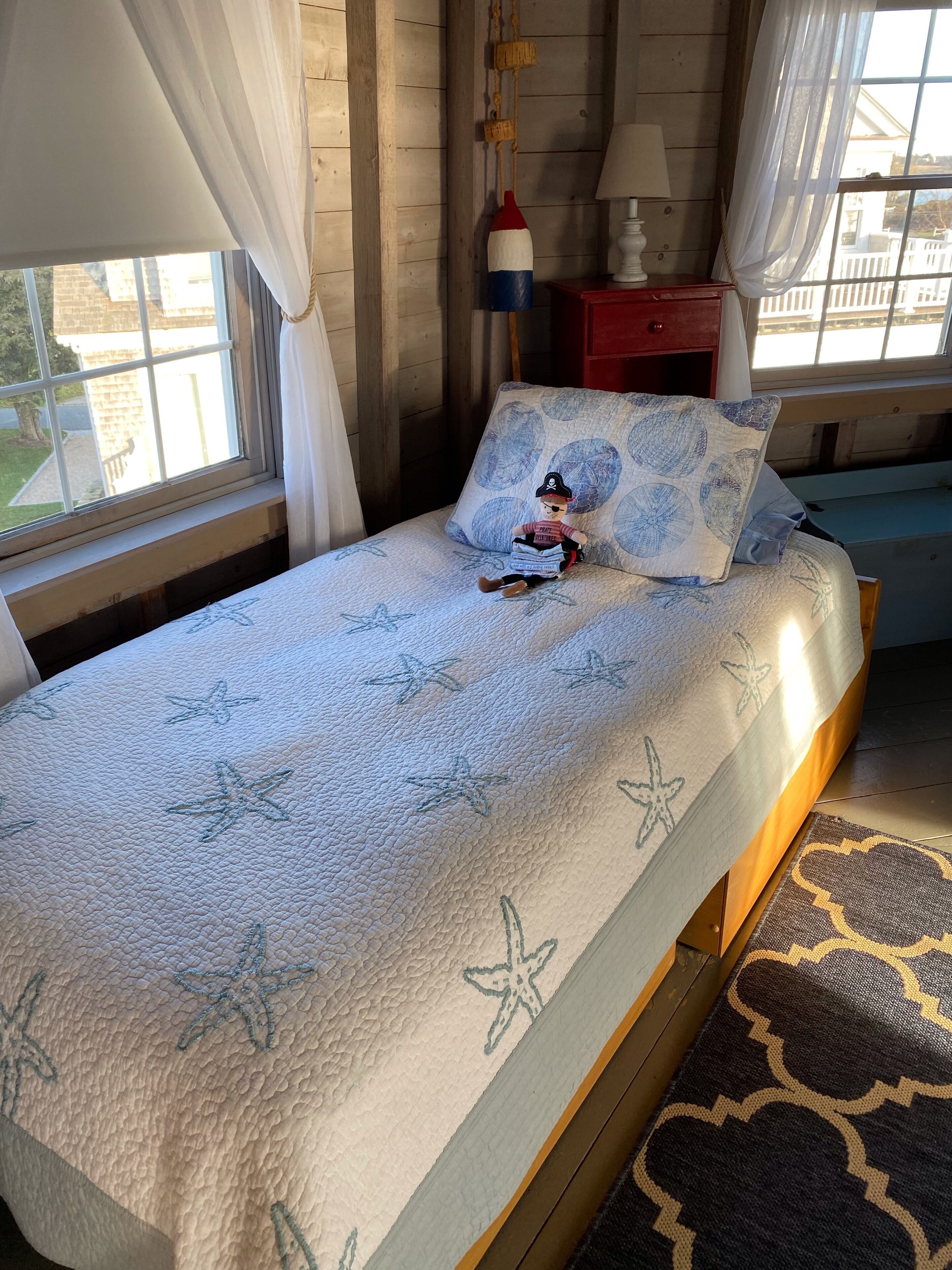 A bed with a starfish-patterned bedspread, a plush toy on the pillow, and a view of the outside through a window with curtains.
