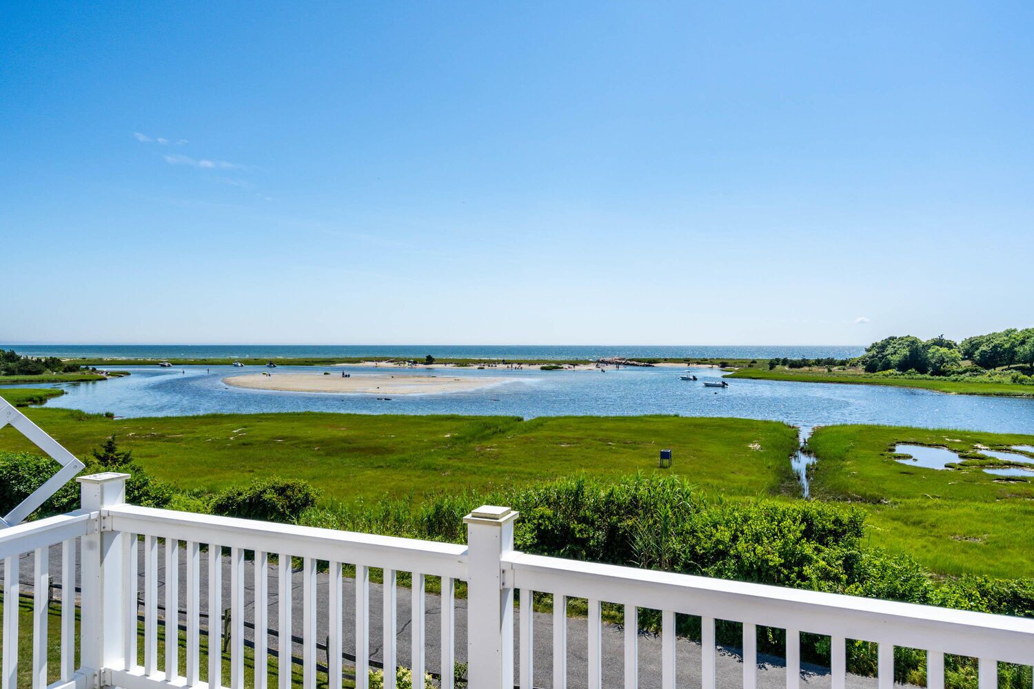 A view of a river with a sandy beach and grassy areas, a clear blue sky, and a white fence in the foreground.