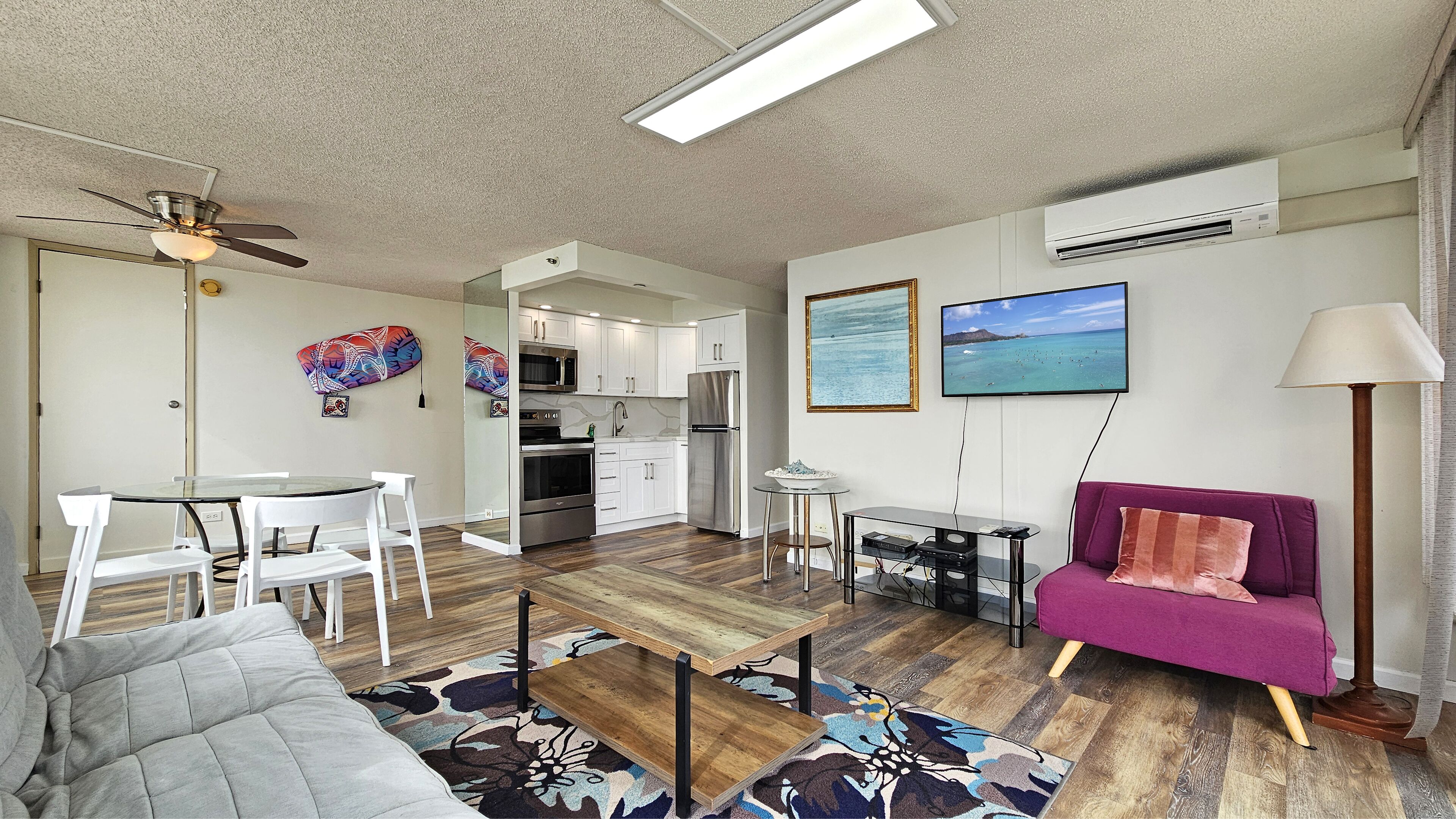 Un salon moderne avec un canapé, une table basse et un coin repas. La cuisine est dotée de meubles blancs et d’appareils électroménagers en acier inoxydable. On peut voir un ventilateur de plafond et un appareil de climatisation.