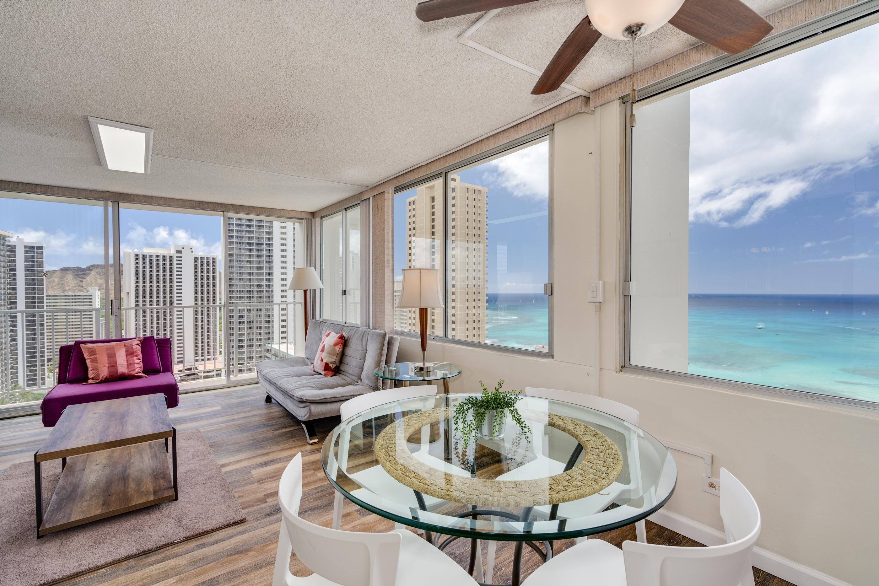 Glass-top dining table with white chairs beside floor-to-ceiling windows overlooking ocean and cityscape.