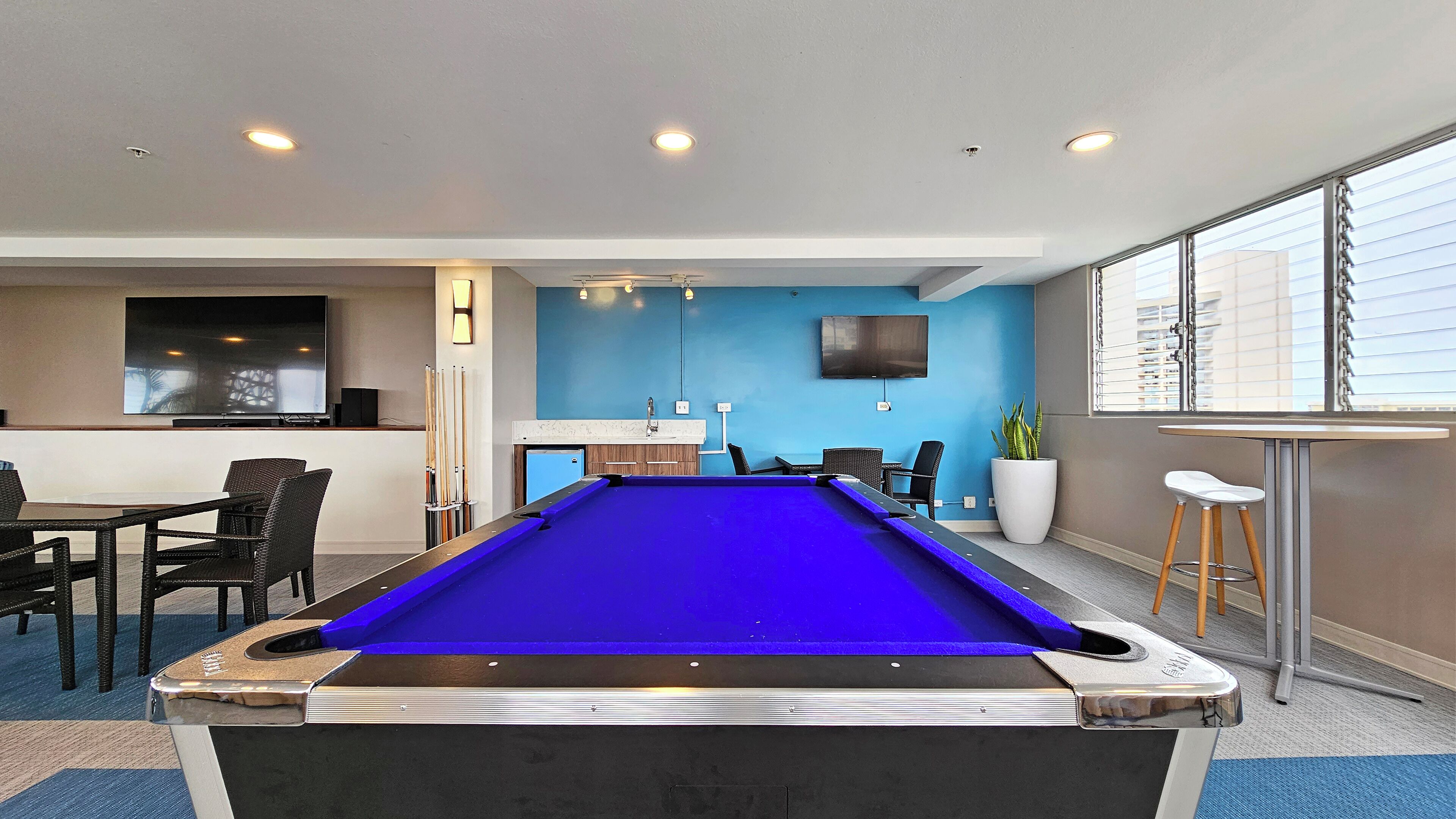 Una mesa de billar en una sala con pared azul, un televisor montado y una zona de bar con taburetes.