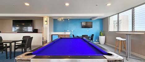 Una mesa de billar en una sala con pared azul, un televisor montado y una zona de bar con taburetes.