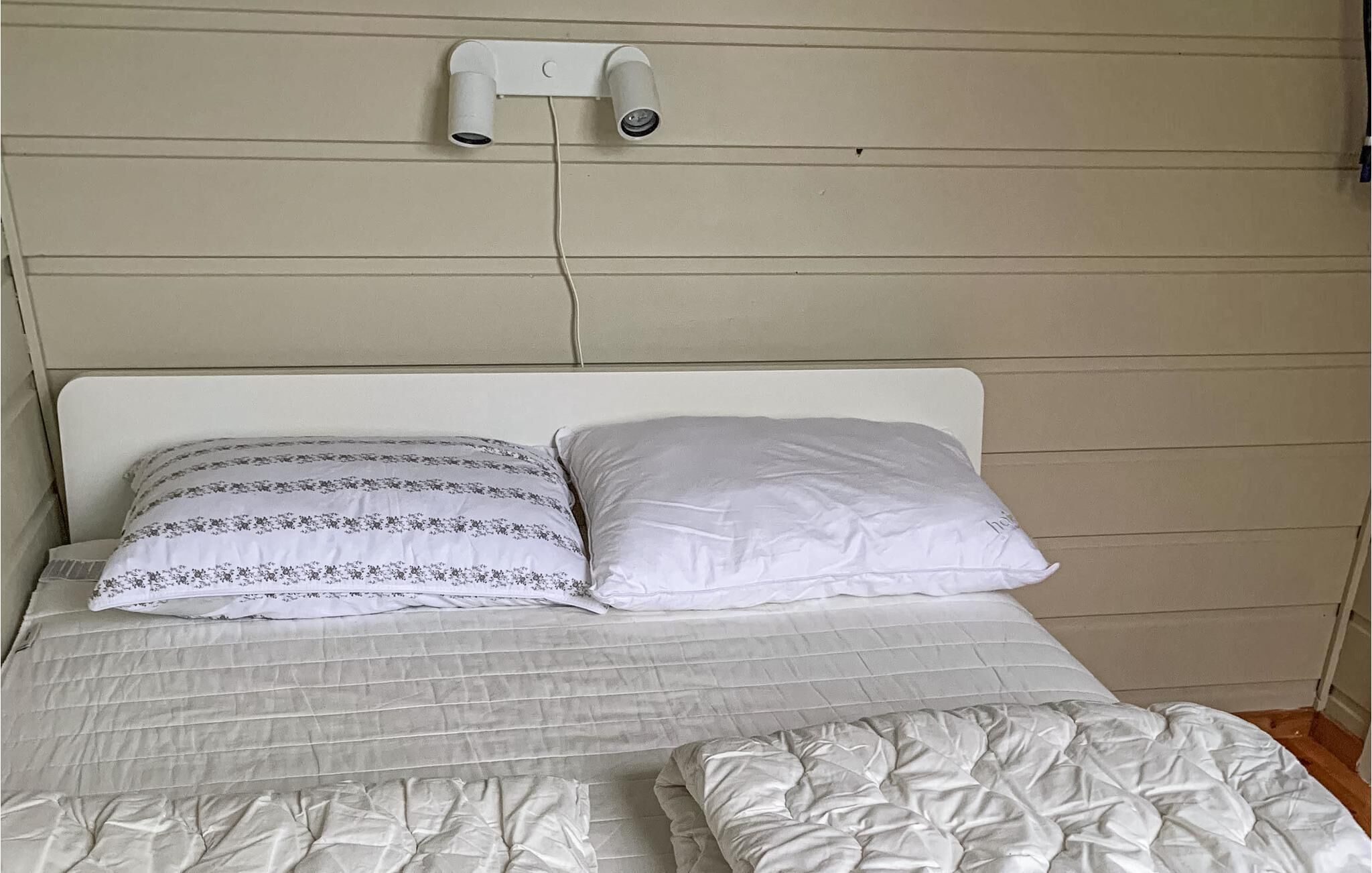 Un lit avec deux oreillers, une tête de lit blanche et une caméra de surveillance fixée au mur.
