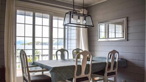 Un comedor con una mesa y sillas, ventanas amplias con vista al mar, una lámpara colgante y un espejo en la pared.