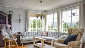Una sala de estar acogedora con un sofá gris, una mesa de centro de madera y un sillón estampado. El suelo es de madera, hay una planta colgante y una ventana con vistas a la vegetación.