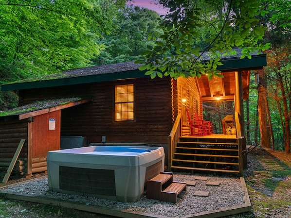A log cabin with a hot tub, wooden stairs, and a covered area with red chairs.