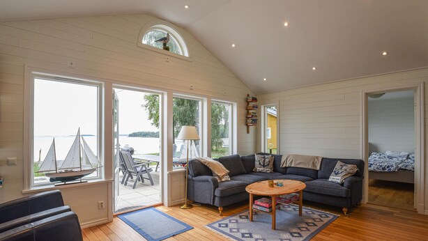 Ein geräumiges Wohnzimmer mit großem Fenster mit Blick auf ein Gewässer, eine gemütliche Sitzecke mit Sofa und Couchtisch sowie ein kleines Segelbootmodell auf der Fensterbank.