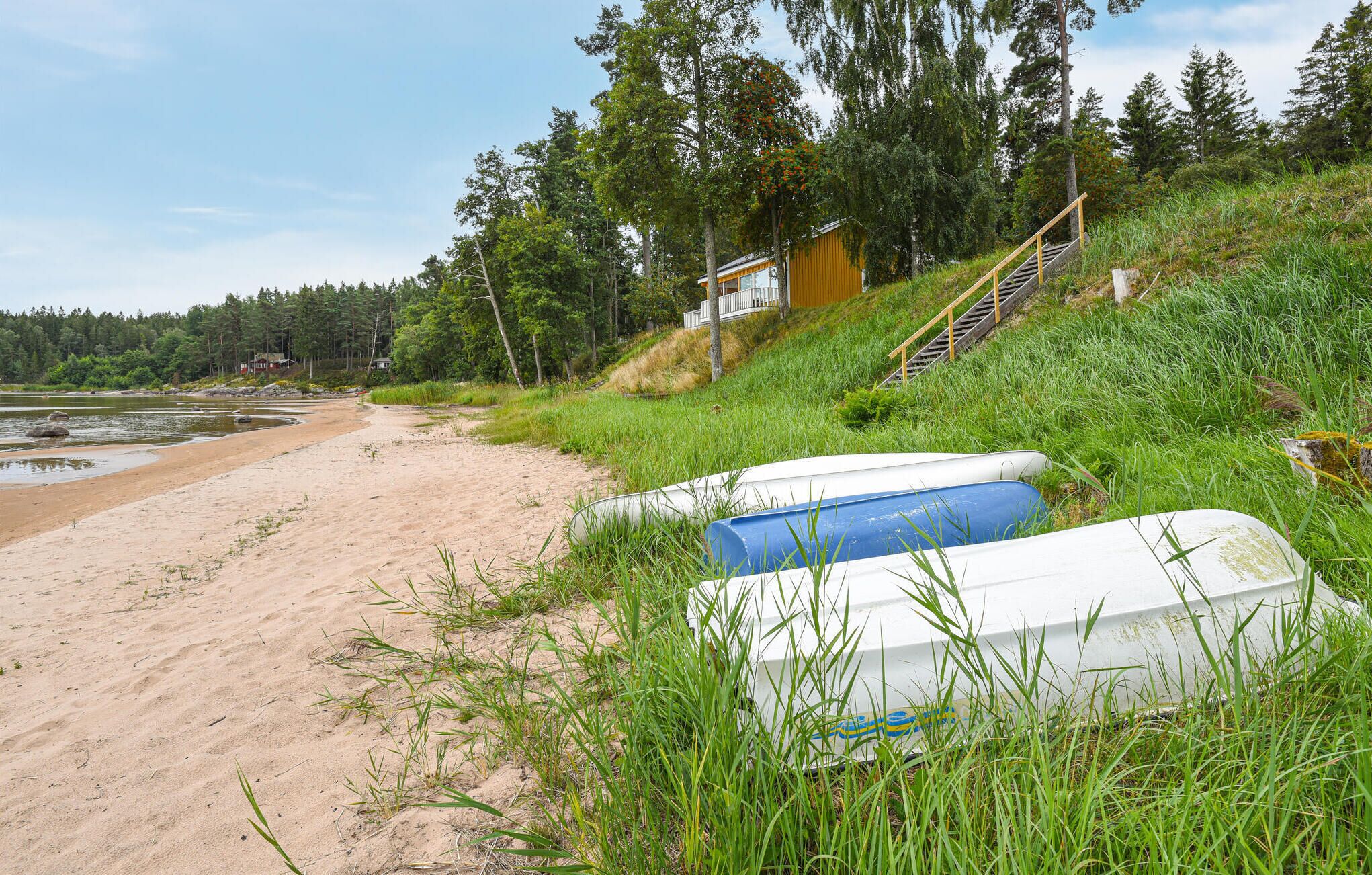 A beach with a small cabin and a staircase leading up to it. There are two boats on the grass near the cabin.