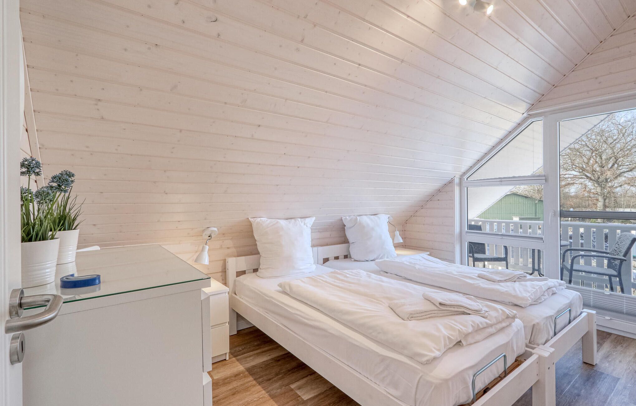Ein Schlafzimmer mit Holzboden, einem Bett mit weißer Bettwäsche, einem kleinen Nachttisch und einem Fenster mit Blick nach draußen.