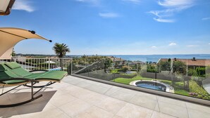 Uma varanda com vista para uma piscina, uma palmeira e um corpo d'água ao longe.