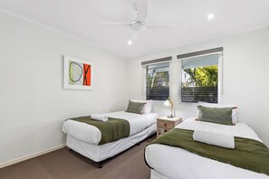 A hotel room with two single beds, a ceiling fan, a window with blinds, and a painting on the wall.