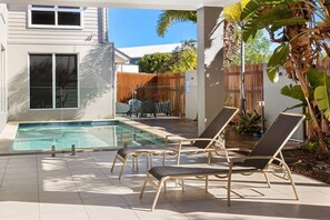 En pool med solstolar, ett trästaket och ett hus i bakgrunden.