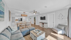 Un salon moderne avec un canapé, une table basse et un téléviseur. On y trouve également un coin repas avec une table et des chaises. Les murs sont blancs et la pièce est équipée d’un ventilateur de plafond.
