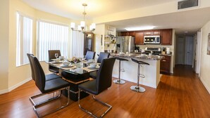 A modern kitchen with stainless steel appliances, a dining area with a glass table and black chairs, and wooden flooring.