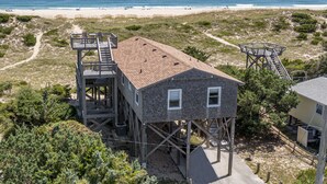 Ett hus vid stranden med en träveranda och en trappa som leder upp till ett andra våningsplan, med utsikt över stranden.