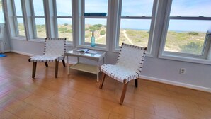 Ett rum med två fåtöljer i rotting, ett litet bord och stora fönster med utsikt över en strand och sanddyner.
