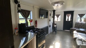 Una cocina con estufa, repisas con ollas, un extintor y una zona de comedor con sofá y televisor.