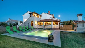 Uma casa de dois andares com piscina, espreguiçadeiras verdes e um espaço de varanda bem iluminado.