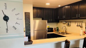 A kitchen with dark wooden cabinets, a stainless steel refrigerator, a chalkboard menu, and a wall clock. - Recently Remodeled, Modern, Nice Condo in a Great Location (Kailua Kona)