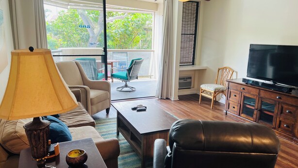 A living room with a sofa, a chair, a coffee table, a TV, and a lamp. There is a balcony with a chair and a table. - Recently Remodeled, Modern, Nice Condo in a Great Location (Kailua Kona)