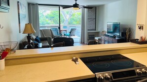 A kitchen with a stove, a dining area with chairs, a TV, and a view of greenery outside. - Recently Remodeled, Modern, Nice Condo in a Great Location (Kailua Kona)