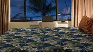 Une chambre avec une grande fenêtre, un lit recouvert d’une couvre-lit à motifs floraux, un ventilateur de plafond et une vue sur l’océan.