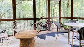 A covered patio with wicker furniture, a bicycle, and a view of a forest. - Cozy Athens Home on the Brook (Athens)