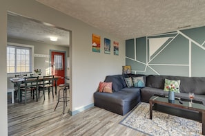 A modern living room with a sectional sofa, a dining area with a wooden table and chairs, and a kitchen visible in the background.