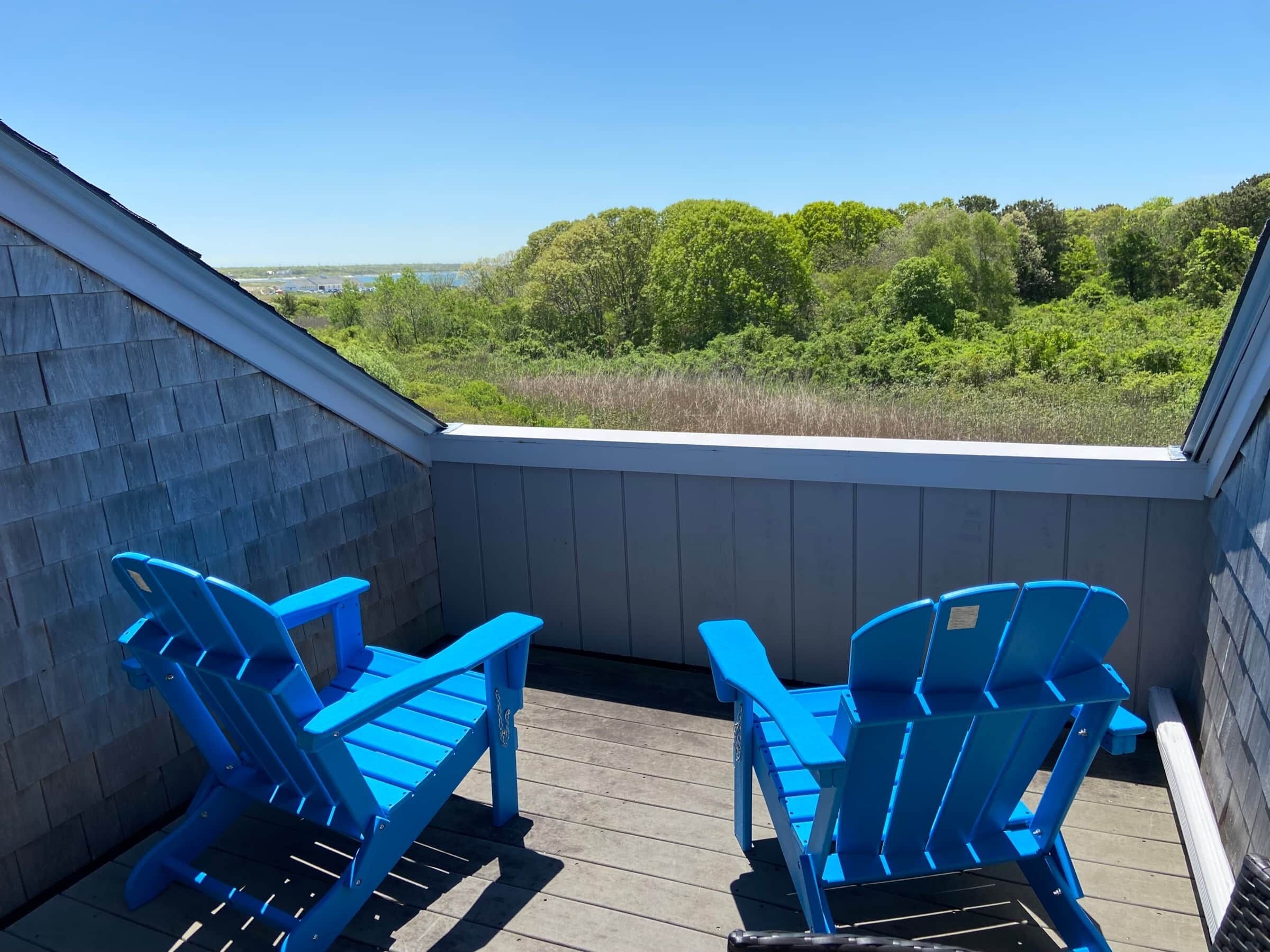 A balcony with two blue Adirondack chairs, wooden deck, and a view of a forested area.