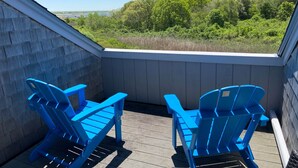 A balcony with two blue Adirondack chairs, wooden deck, and a view of a forested area. - 46 by the Sea, steps to private beach & heated pool, 1-mile from Main St., ocean views,  AC, & Wi-Fi (Barnstable)