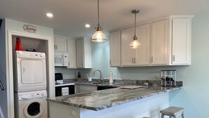 A kitchen with a built-in washer and dryer, a granite countertop, white cabinets, and pendant lighting. - 46 by the Sea, steps to private beach & heated pool, 1-mile from Main St., ocean views,  AC, & Wi-Fi (Barnstable)