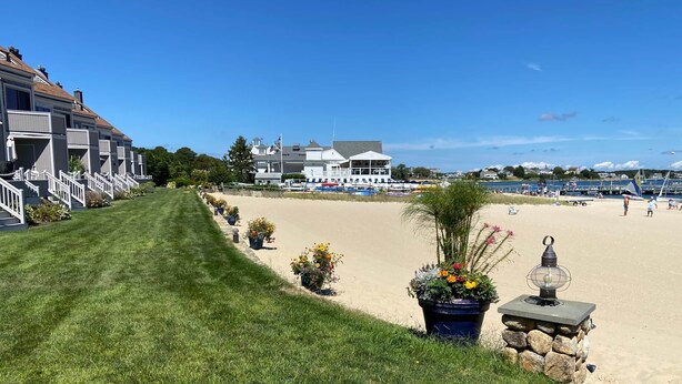 A well-maintained lawn leading to a sandy beach area with potted plants and a stone lamp post. - 46 by the Sea, steps to private beach & heated pool, 1-mile from Main St., ocean views,  AC, & Wi-Fi (Barnstable)