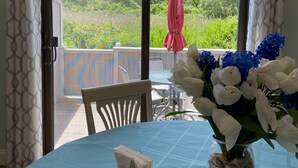 A room with a table set for dining, a vase with flowers, and a view of greenery through a glass door. - 46 by the Sea, steps to private beach & heated pool, 1-mile from Main St., ocean views,  AC, & Wi-Fi (Barnstable)