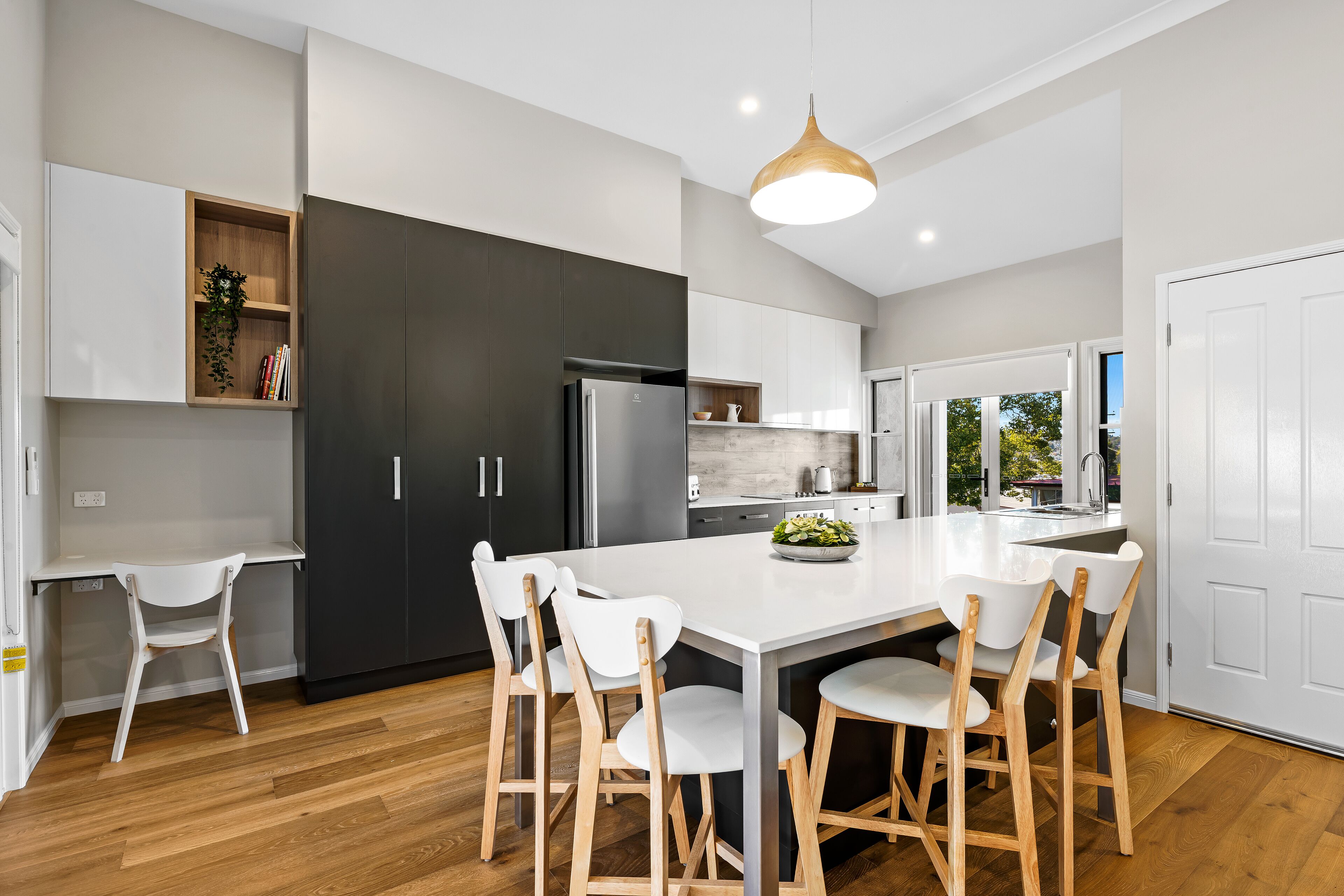 A modern kitchen with a central island, white chairs, dark cabinets, and a wooden ceiling light.