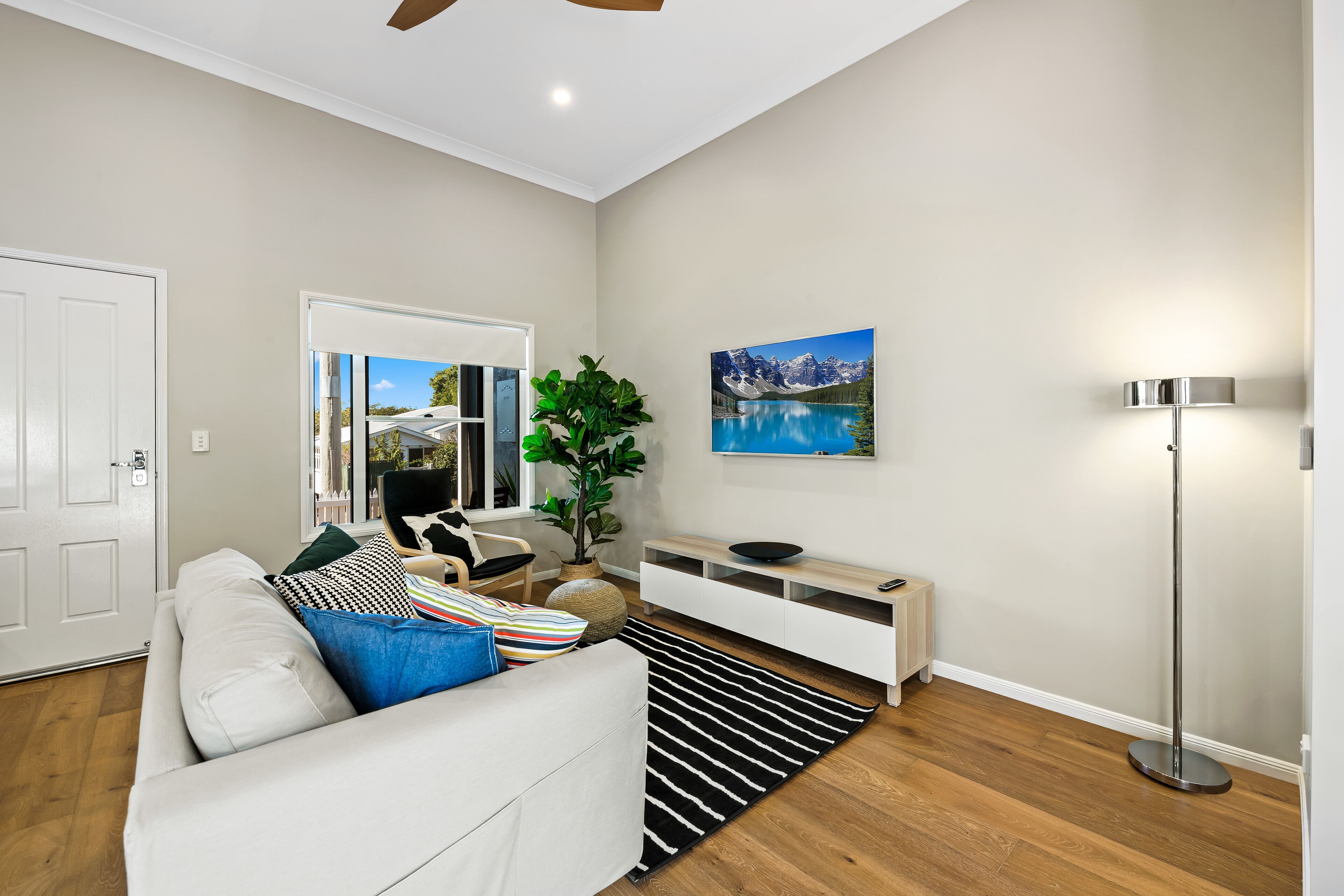 A modern living room with a white sofa, a black and white striped rug, a plant, a painting of a lake, and a floor lamp.