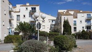 A residential building with multiple balconies, a paved courtyard, and a car parked in front. - Les Villageoises by Interhome (Le Barcarès)