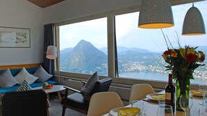 Un comedor con una mesa preparada para dos, un sofá con cojines estampados y un ventanal con vistas a una montaña y una ciudad.