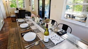 Comedor con una larga mesa de madera dispuesta para seis personas, con platos blancos, cubiertos y una botella de vino.