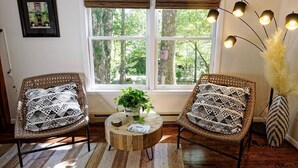 Una habitación con dos sillones de mimbre con cojines estampados, una mesa de centro redonda de madera con una planta y un ventanal con persianas.