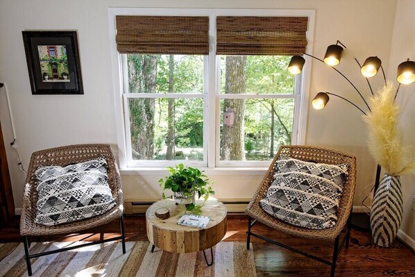 Una habitación con dos sillones de mimbre con cojines estampados, una mesa de centro redonda de madera con una planta y un ventanal con persianas.