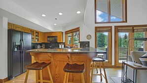 A modern kitchen with wooden cabinets, a central island with stools, a refrigerator, and a dining area with a sofa and table.