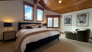A bedroom with a large bed, two windows with red valances, a ceiling with wooden beams, and two framed pictures on the wall.