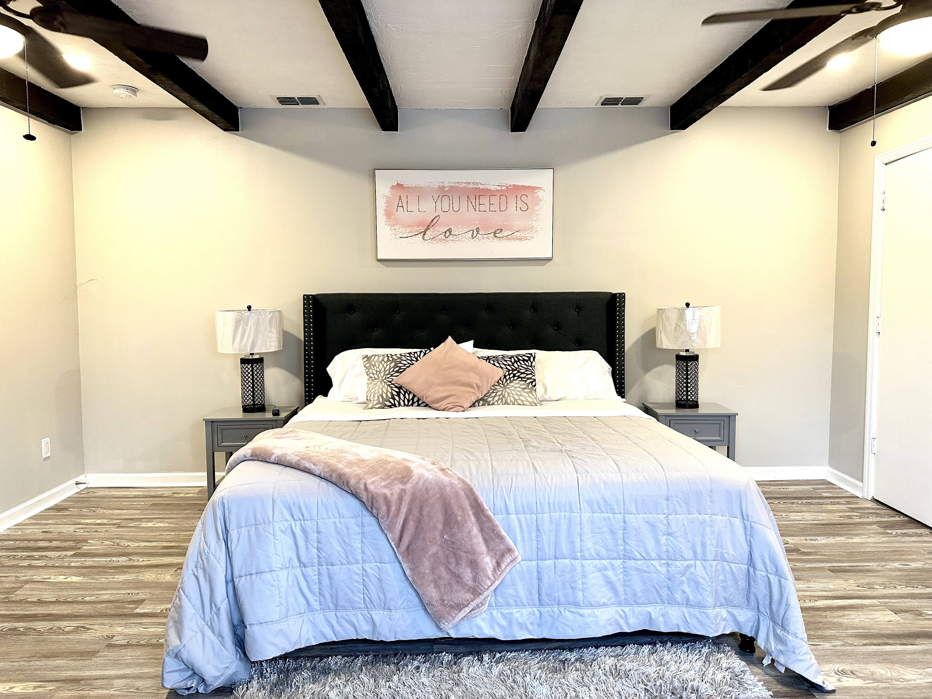 A bedroom with a large bed, two bedside tables with lamps, a ceiling fan, and a motivational quote on the wall.